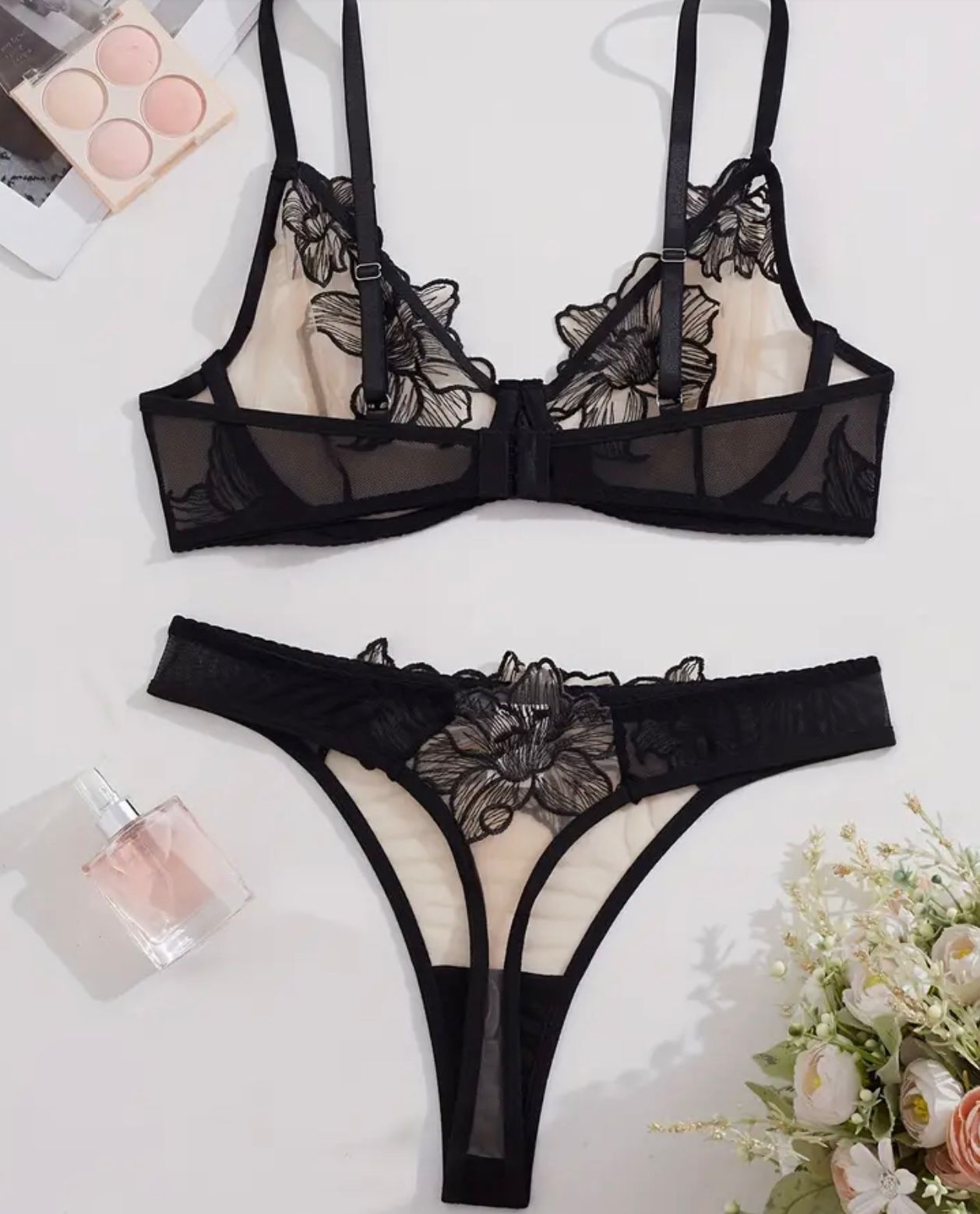 Black lace lingerie set on a white background with a small bouquet of flowers and perfume bottle.