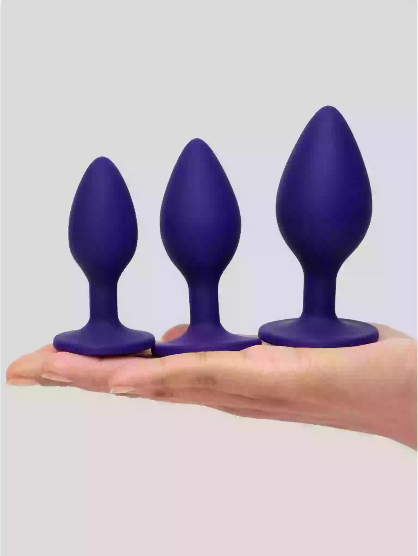 Three purple silicone butt plugs of different sizes held in a hand against a light gray background