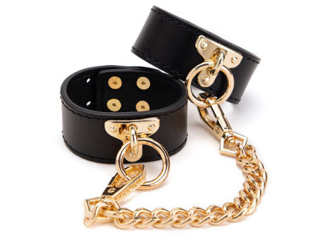 Black leather cuffs with gold hardware and chain on a white background